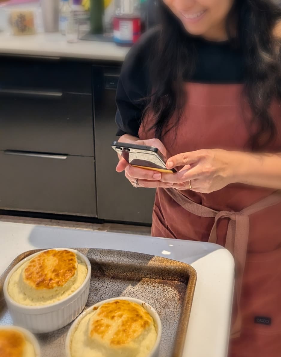 A phone propped in a kitchen showing a cooking video, with a finished home-cooked dish next to it.